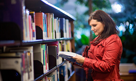 A woman in a red jacket reading a book in a library aisle.