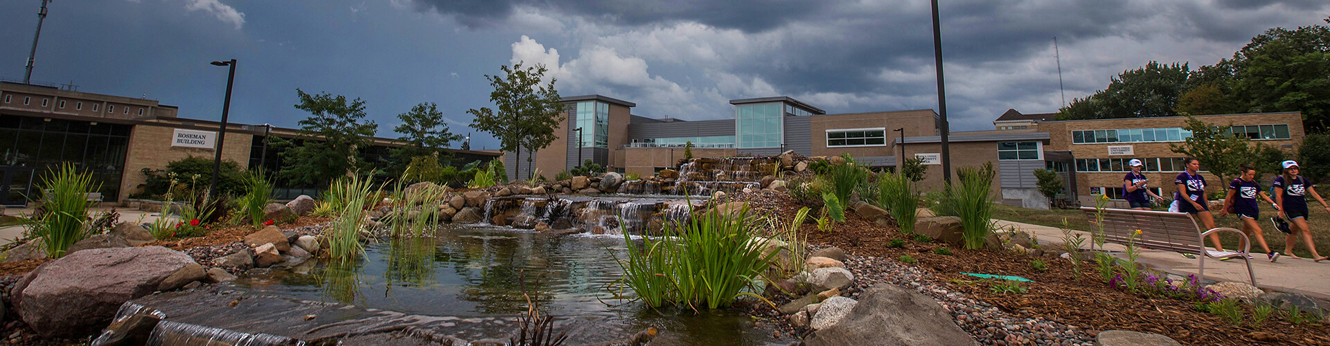 A landscaped water feature with cascading falls in front of a modern building, under a partly cloudy sky, with several people walking along the path.