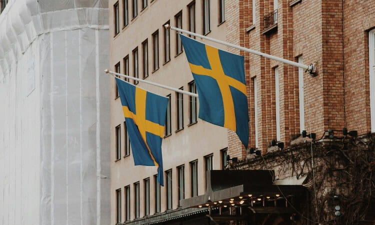 Two Swedish flags fluttering from white poles on the brick facade of an urban building.